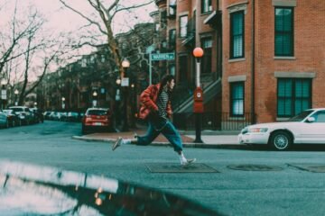 Hotfoot meaning shown by a young man sprinting across a city street in a red jacket